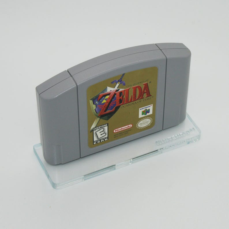 May include: A gray Nintendo 64 game cartridge for The Legend of Zelda: Ocarina of Time. The cartridge is on a clear plastic stand. The game is rated E for Everyone by the ESRB.