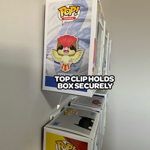 May include: A white wall mount display with a white clip holding a Pop! Games Pidgeotto figure in a box. The text "TOP CLIP HOLDS BOX SECURELY" is printed on the wall mount.
