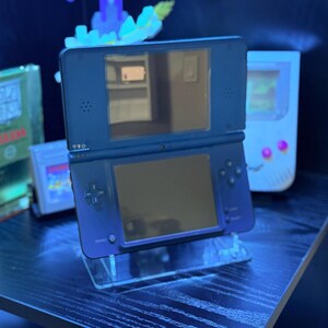 May include: A dark blue Nintendo DS handheld game console, open and displayed on a clear acrylic stand. The console features two screens, a directional pad, and action buttons. Other retro gaming consoles are visible in the background.