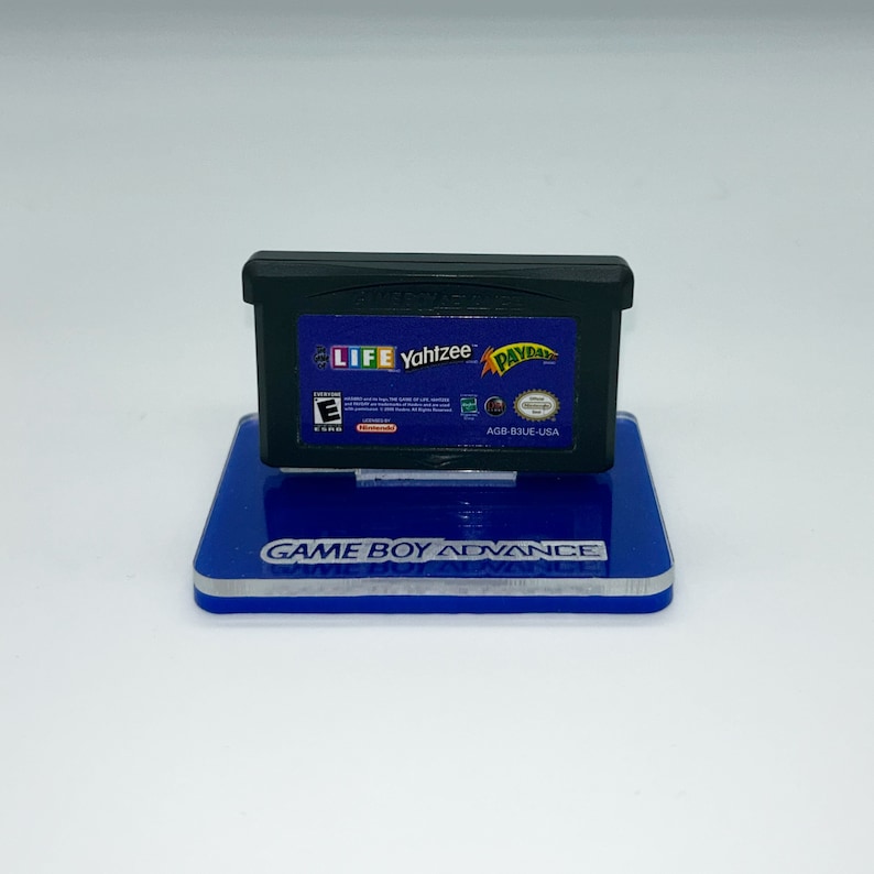 May include: A black Game Boy Advance game cartridge with a blue and yellow label. The label features the text "LIFE Yahtzee" and "PAYDAY". The game is rated "E" for Everyone by the ESRB. The cartridge is on a blue display stand with the text "GAME BOY ADVANCE".
