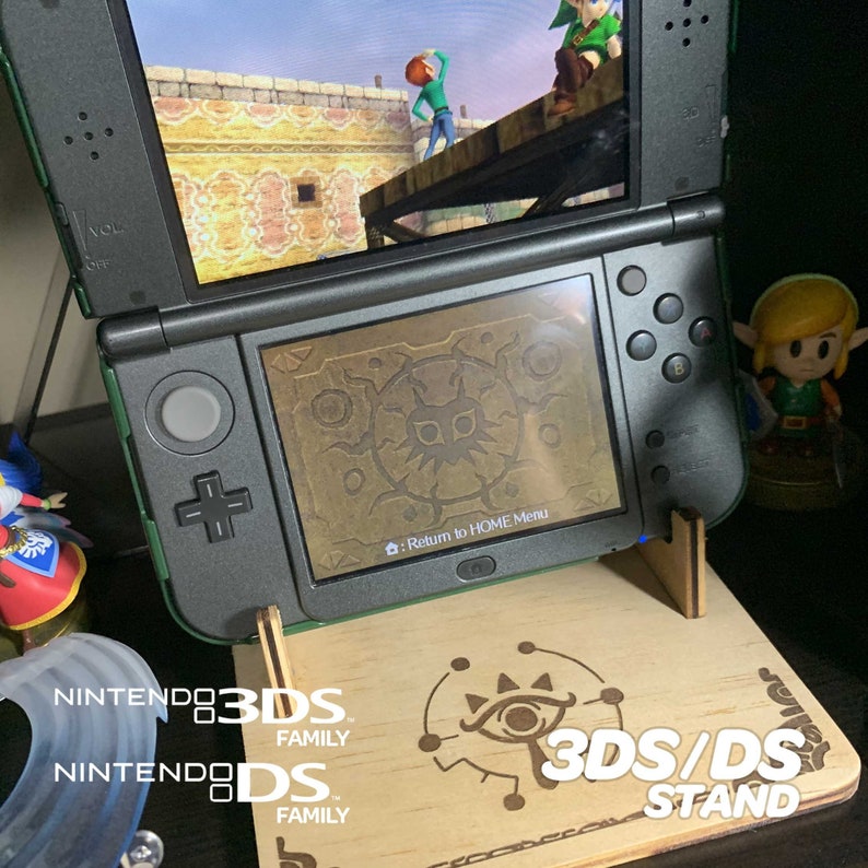 May include: A wooden stand for a Nintendo 3DS or DS handheld gaming console. The stand is engraved with the words "3DS/DS STAND" and a stylized eye design. The stand is shown with a green and black Nintendo 3DS console on top of it.