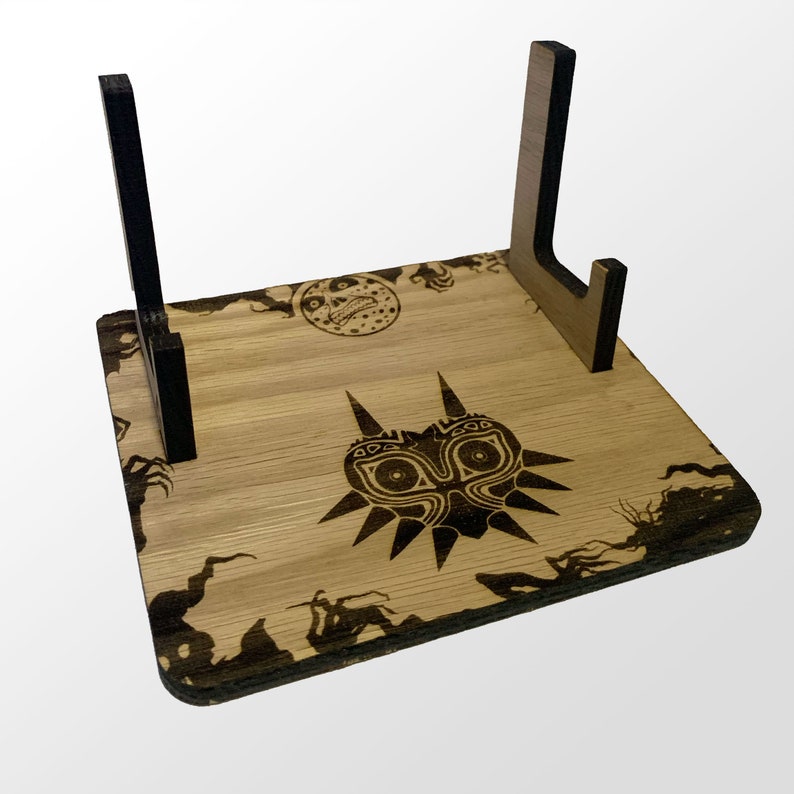 May include: A wooden stand with a black and white design of the Majora's Mask from the Legend of Zelda video game series. The stand has two black vertical supports and a base with a brown wood grain finish.