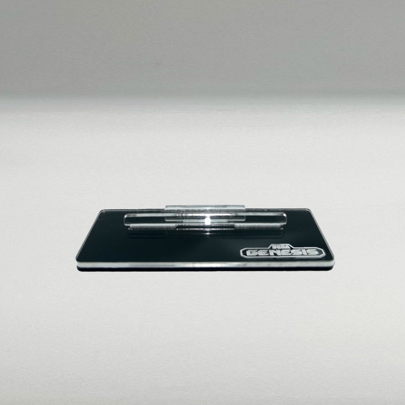 Op de afbeelding: Een zwarte acrylstandaard met een heldere acrylstaaf om een Sega Genesis-spelcartridge te presenteren. De standaard heeft het Sega Genesis-logo erop gedrukt.