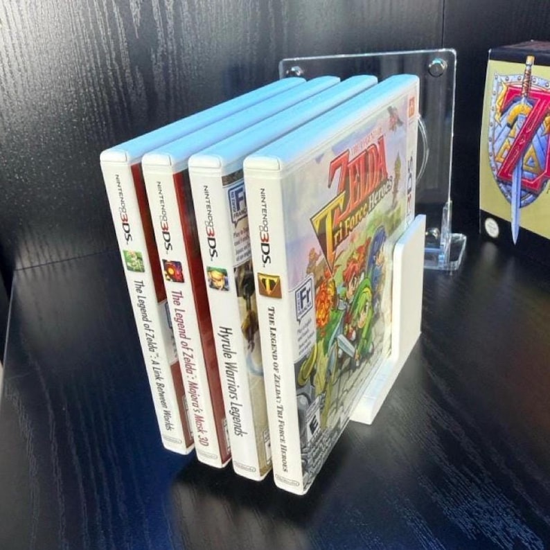 Puede incluir: Un expositor blanco que contiene cuatro carcasas de juegos de Nintendo 3DS. Los juegos son "The Legend of Zelda: A Link Between Worlds", "The Legend of Zelda: Majora's Mask 3D", "Hyrule Warriors Legends" y "The Legend of Zelda: Tri Force Heroes".