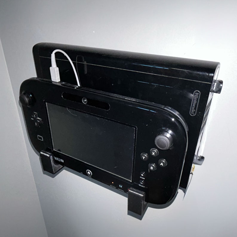 May include: A black Nintendo Wii U console and a black Wii U gamepad are mounted on a wall. The console is plugged in with a white cord. The gamepad has a black screen and buttons.