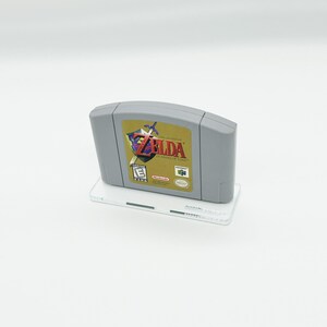 May include: A gray Nintendo 64 game cartridge with a gold label featuring the text "The Legend of Zelda: Ocarina of Time" and an image of Link holding a sword. The game is rated "E" for Everyone by the ESRB.