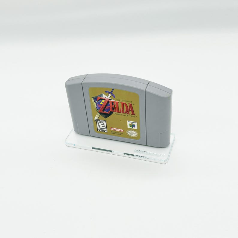 May include: A gray Nintendo 64 game cartridge with a gold label featuring the text "The Legend of Zelda: Ocarina of Time" and an image of Link holding a sword. The game is rated "E" for Everyone by the ESRB.