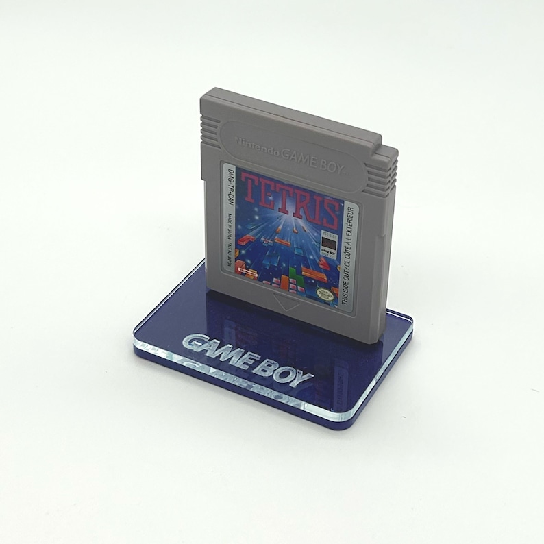 May include: A gray Game Boy cartridge for the game Tetris is displayed on a blue acrylic stand with the words "GAME BOY" printed on it.