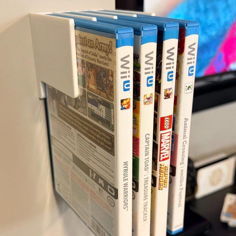 May include: A white wall-mounted game holder with four Wii U game cases. The game cases have blue tops and white spines with game titles like "Hyrule Warriors", "Captain Toad: Treasure Tracker", and "LEGO Marvel Super Heroes".