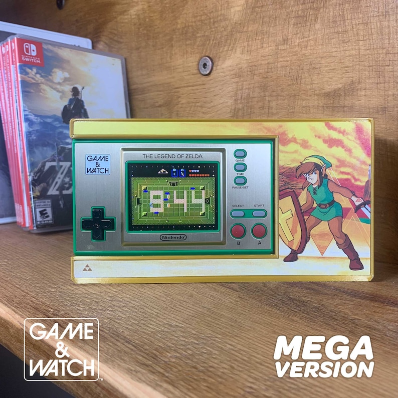 May include: A yellow and green Game & Watch handheld video game console featuring The Legend of Zelda. The screen displays the game title and a pixelated image of Link, the main character, with the words "9:44" on the screen. The console has a green and yellow color scheme with a gold trim. The console is on a wooden surface.