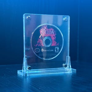 May include: A clear acrylic display stand holding a Zelda GameCube disc. The disc features the game's title and logo against a fiery background. The stand has a rectangular base and is secured with small screws.