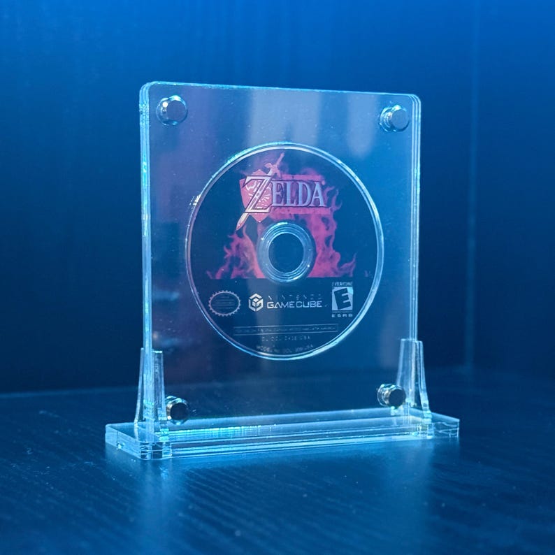 May include: A clear acrylic display stand holding a Zelda GameCube disc. The disc features the game's title and logo against a fiery background. The stand has a rectangular base and is secured with small screws.