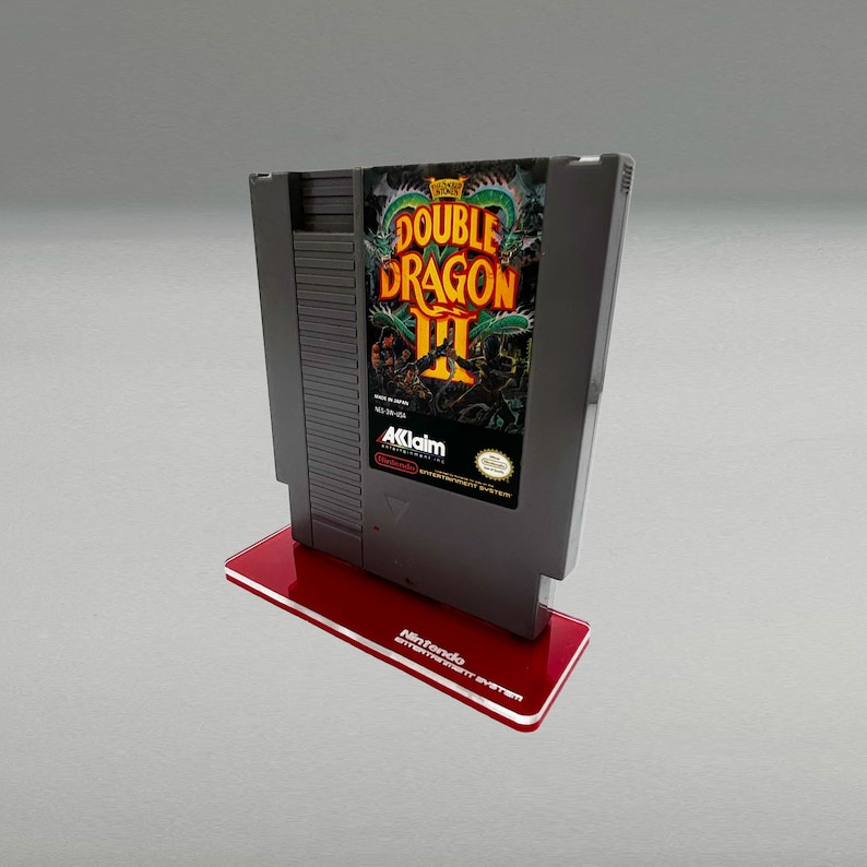 May include: A gray Nintendo Entertainment System game cartridge for Double Dragon III, featuring a green, red, and black illustration of a dragon on the front. The cartridge is displayed on a red acrylic stand.