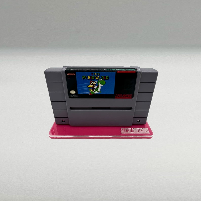 May include: A gray Super Nintendo game cartridge for Super Mario World, displayed on a pink stand with the words "SUPER NINTENDO" printed on the front.