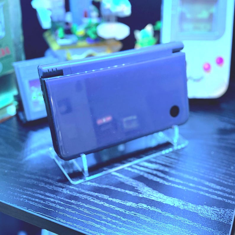 May include: A dark blue Nintendo DS Lite handheld game console, displayed on a clear acrylic stand. The console is closed, showing its smooth, reflective surface. Other retro gaming devices are visible in the background.