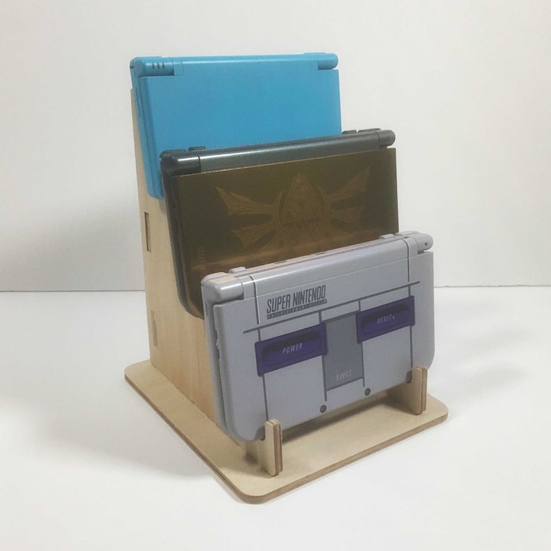 May include: A wooden stand with three slots for video game consoles. The top slot holds a blue Nintendo DS, the middle slot holds a gold Nintendo 3DS, and the bottom slot holds a gray Super Nintendo.