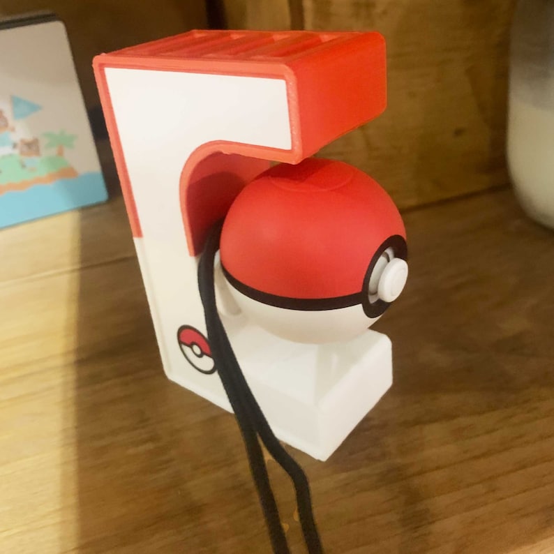 May include: A white and red Pokeball holder with a black strap. The Pokeball is red and white with a black ring around the middle. The holder has a small Pokeball logo on the side.