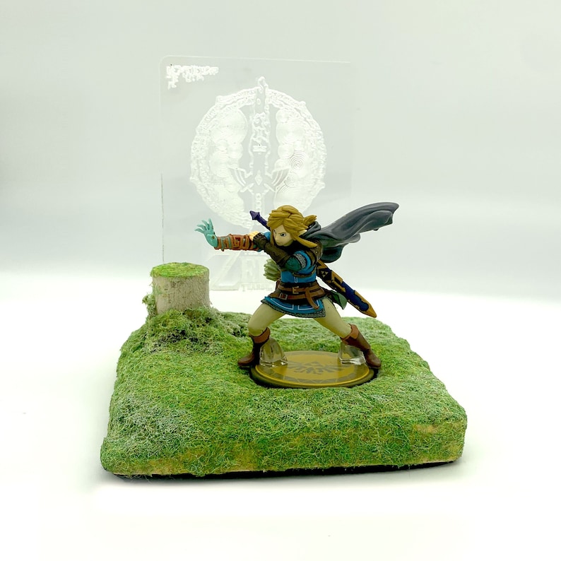 May include: A detailed figurine of a character from The Legend of Zelda, posed on a grassy base with a tree stump. The figure is wearing a blue and brown outfit, with a cape and sword. A clear acrylic panel with a design is in the background.