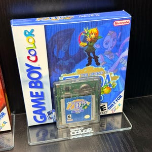 May include: A Game Boy Color video game cartridge and box set for "The Legend of Zelda: Oracle of Ages." The box is blue with a character illustration and the Nintendo logo. The cartridge is in a clear plastic case.
