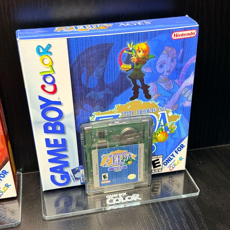 May include: A Game Boy Color video game cartridge and box set for "The Legend of Zelda: Oracle of Ages." The box is blue with a character illustration and the Nintendo logo. The cartridge is in a clear plastic case.