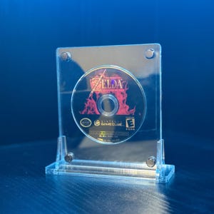 May include: A clear acrylic display case holding a Zelda video game disc. The disc features the game's title and a fiery graphic. The case has a rectangular shape with a base and is secured with small screws.