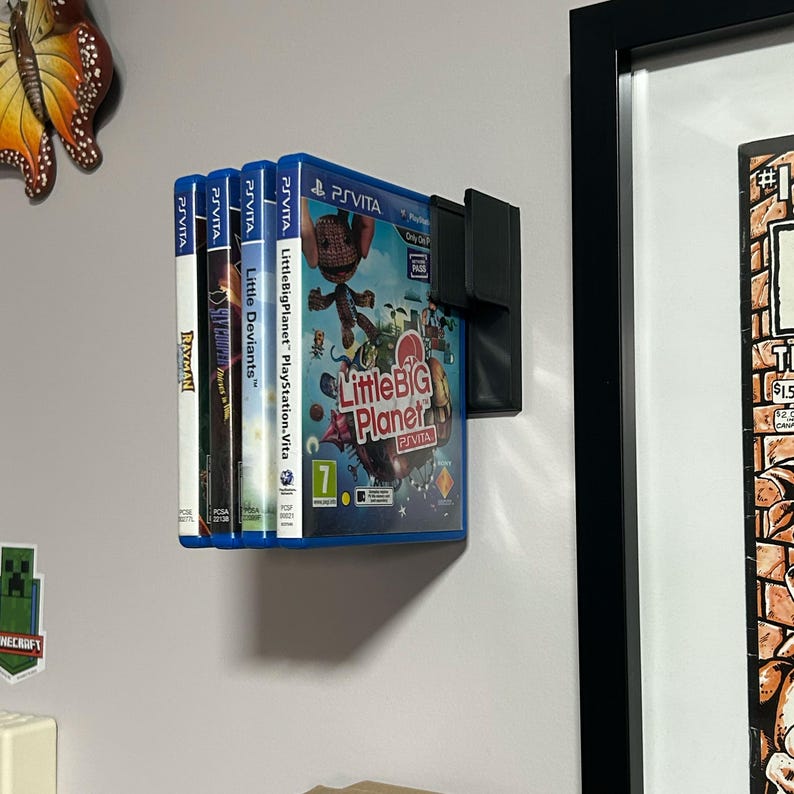 May include: A wall-mounted video game storage rack with four blue video game cartridges. The cartridges are for the PlayStation Vita and include the games LittleBigPlanet, Little Deviants, and Rayman.