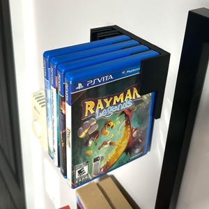 May include: A black wall mount game holder with four video game cases. The game case on the right is for the game Rayman Legends for the Playstation Vita.