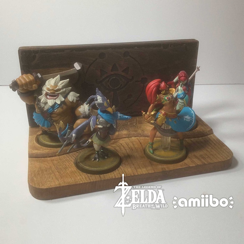 May include: A wooden display stand for amiibo figures from The Legend of Zelda: Breath of the Wild. The stand features a brown wooden base with a cutout for each figure. The stand also features a brown wooden back panel with a carved design of the Triforce and the words "The Legend of Zelda: Breath of the Wild". The stand is designed to hold four amiibo figures: a blue and purple bird-like figure, a red-haired female figure, a brown and white figure with a sword, and a red-haired female figure with a blue shield.