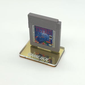 May include: A gray Game Boy cartridge for the game Tetris is displayed on a clear acrylic stand with the words "GAME BOY" printed on the stand. The cartridge has a blue, yellow, and red label with the text "TETRIS" printed in white.