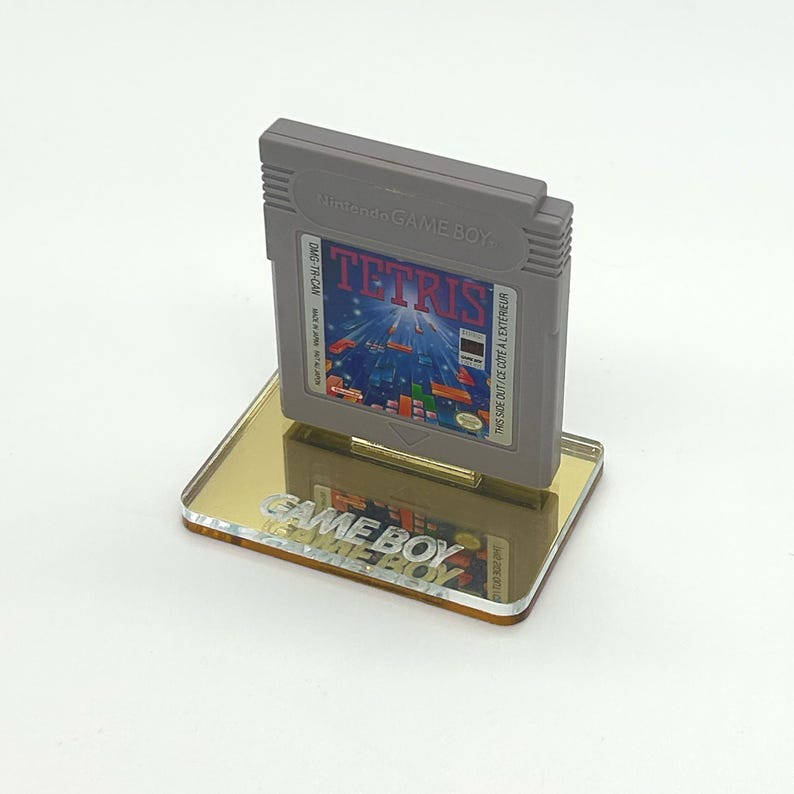 May include: A gray Game Boy cartridge for the game Tetris is displayed on a clear acrylic stand with the words "GAME BOY" printed on the stand. The cartridge has a blue, yellow, and red label with the text "TETRIS" printed in white.