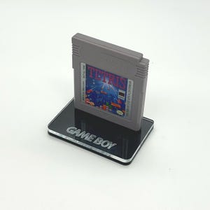 May include: A gray Game Boy cartridge with the text "Nintendo GAME BOY" and "DMG-TR-CAN" on the label. The cartridge is titled "TETRIS" and has a colorful image of falling blocks. The cartridge is displayed on a black acrylic stand with the text "GAME BOY" engraved on the front.