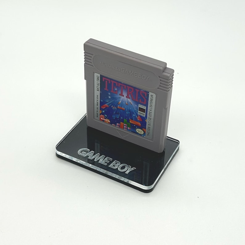 May include: A gray Game Boy cartridge with the text "Nintendo GAME BOY" and "DMG-TR-CAN" on the label. The cartridge is titled "TETRIS" and has a colorful image of falling blocks. The cartridge is displayed on a black acrylic stand with the text "GAME BOY" engraved on the front.