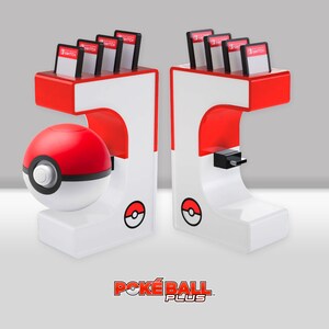 May include: A white and red Poke Ball Plus charging stand with two slots for Nintendo Switch game cartridges. The stand features a Poke Ball design with a red and white Poke Ball on the front. The stand is labeled "POKE BALL PLUS".