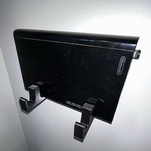 May include: A black Nintendo Wii console mounted on a wall with two black plastic hooks. The console has a black finish and the Nintendo logo is visible on the front.