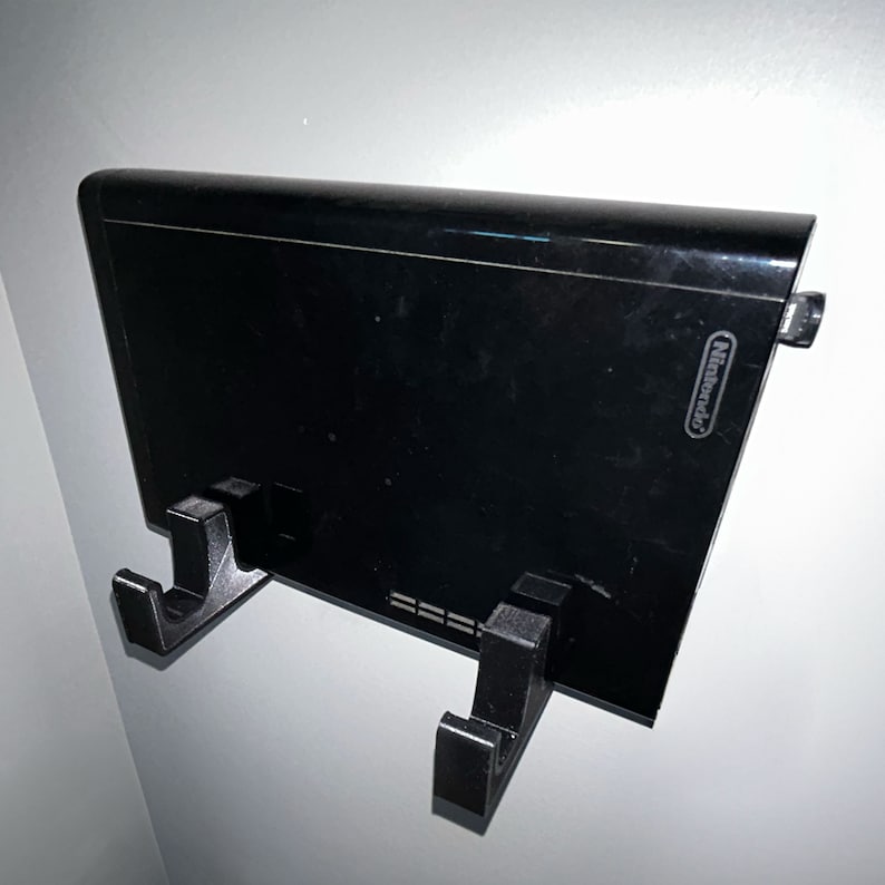 May include: A black Nintendo Wii console mounted on a wall with two black plastic hooks. The console has a black finish and the Nintendo logo is visible on the front.