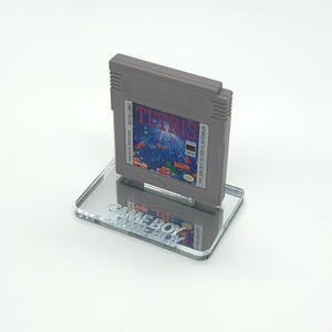 May include: A gray Game Boy cartridge for the game Tetris is displayed on a clear acrylic stand with the words "GAME BOY" printed on it.