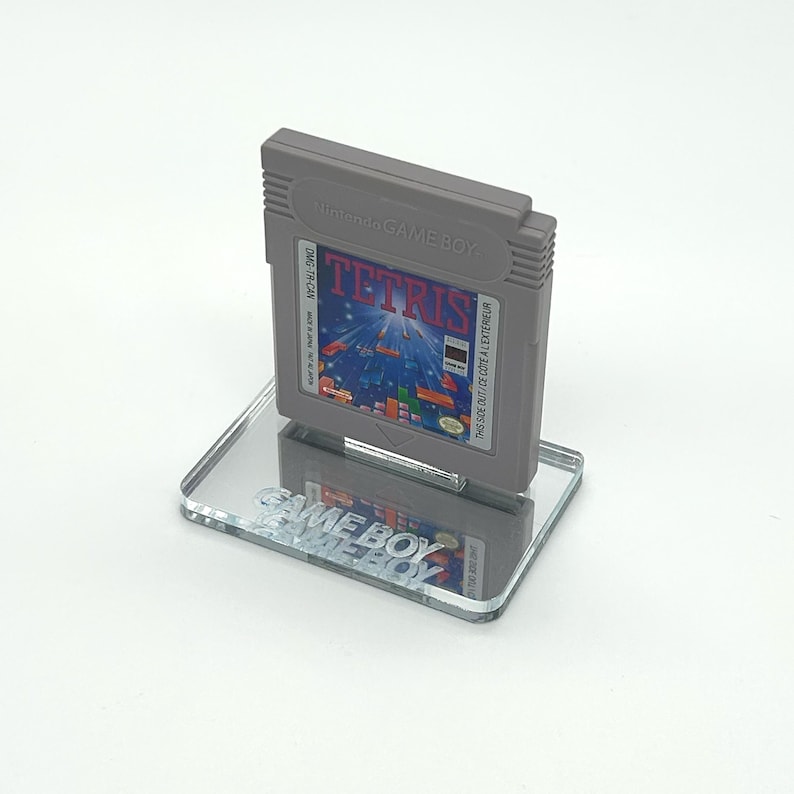 May include: A gray Game Boy cartridge for the game Tetris is displayed on a clear acrylic stand with the words "GAME BOY" printed on it.