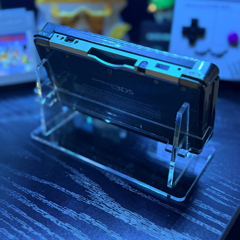 May include: A black Nintendo 3DS handheld game console is displayed on a clear acrylic stand. The console has a silver trim and the words "Nintendo 3DS" are visible. The stand is designed to showcase the device.