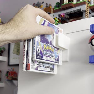 Puede incluir: Un conjunto de cartuchos de videojuegos de Nintendo 3DS, incluyendo "Pokemon Moon" y "Pokemon Ultra Moon", sostenidos en un soporte de pl&aacute;stico blanco. Las cajas de los juegos presentan gr&aacute;ficos coloridos y texto. El soporte est&aacute; montado en una superficie blanca.