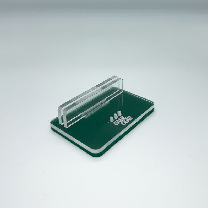 May include: A green acrylic stand with a clear acrylic insert. The stand has the words "GAME GEAR" printed on it.