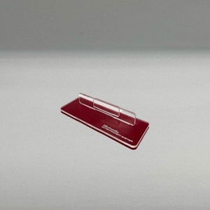 May include: A red acrylic stand for a Nintendo Entertainment System console. The stand has a clear acrylic support for the console and the words "Nintendo Entertainment System" are printed on the base.
