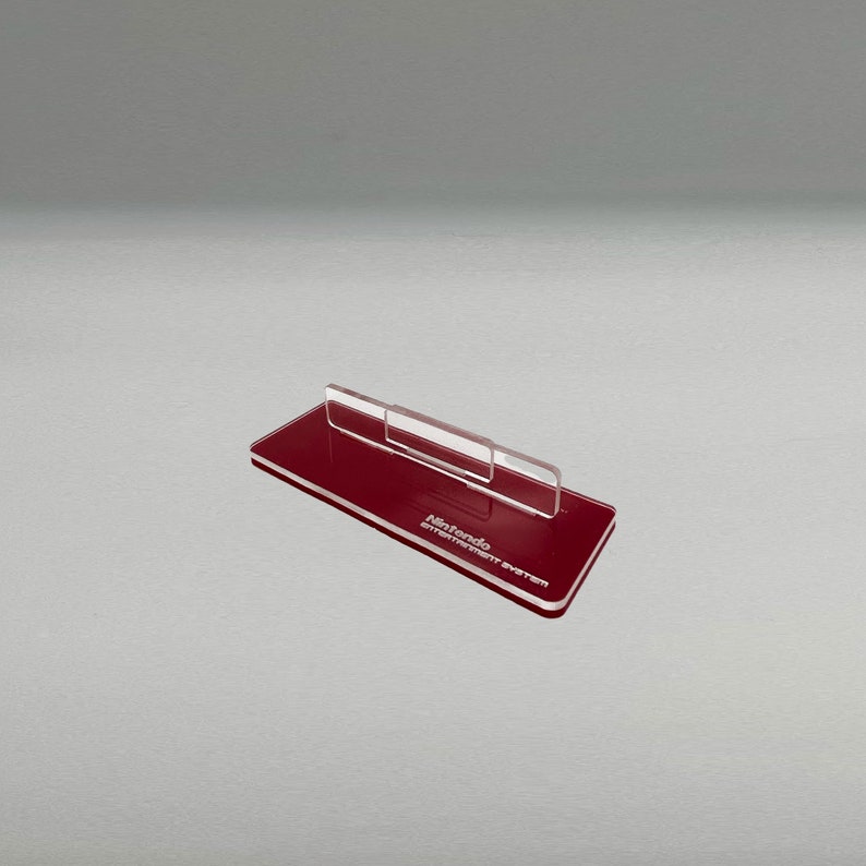 May include: A red acrylic stand for a Nintendo Entertainment System console. The stand has a clear acrylic support for the console and the words "Nintendo Entertainment System" are printed on the base.