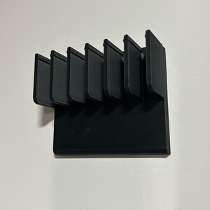 May include: A black, 3D-printed organizer with six vertical slots. The slots are evenly spaced and angled slightly. The organizer is mounted on a white wall. The design is modern and minimalist.