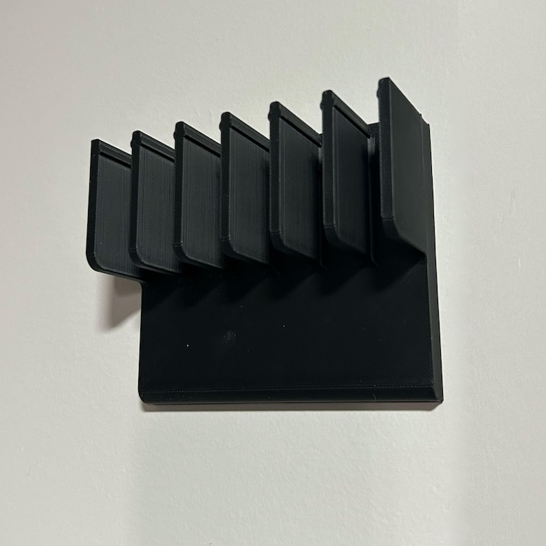 May include: A black, 3D-printed organizer with six vertical slots. The slots are evenly spaced and angled slightly. The organizer is mounted on a white wall. The design is modern and minimalist.