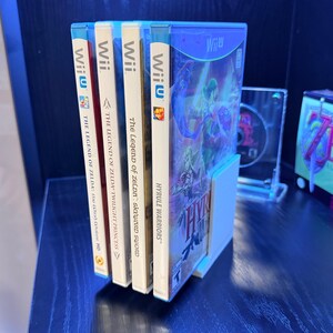 May include: A collection of Wii U video game cases, including "Hyrule Warriors" and "The Legend of Zelda" titles. The game cases are white with blue accents and feature colorful game art. They are held in a white stand.