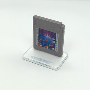May include: A gray Game Boy cartridge with a blue and white Tetris game label. The cartridge is sitting on a clear acrylic stand with the words "Game Boy" printed on it.