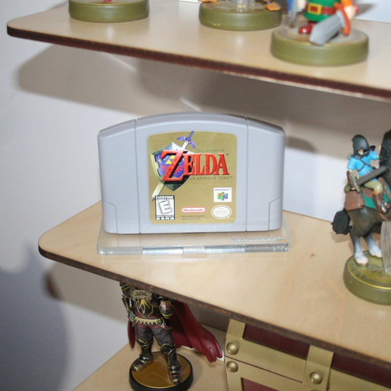 May include: A gray Nintendo 64 game cartridge with a gold label. The label features the text "The Legend of Zelda: Ocarina of Time" and a sword and shield design. The game is rated "E" for Everyone by the ESRB.