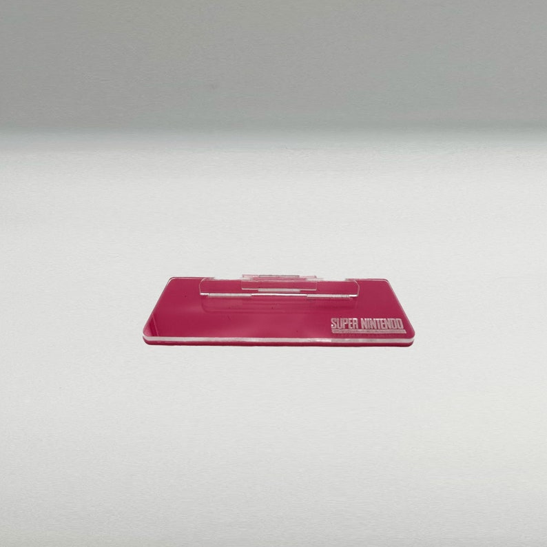 May include: A pink plastic stand for a Super Nintendo game cartridge. The stand has a slot for the cartridge and the words "SUPER NINTENDO" are printed on the front.