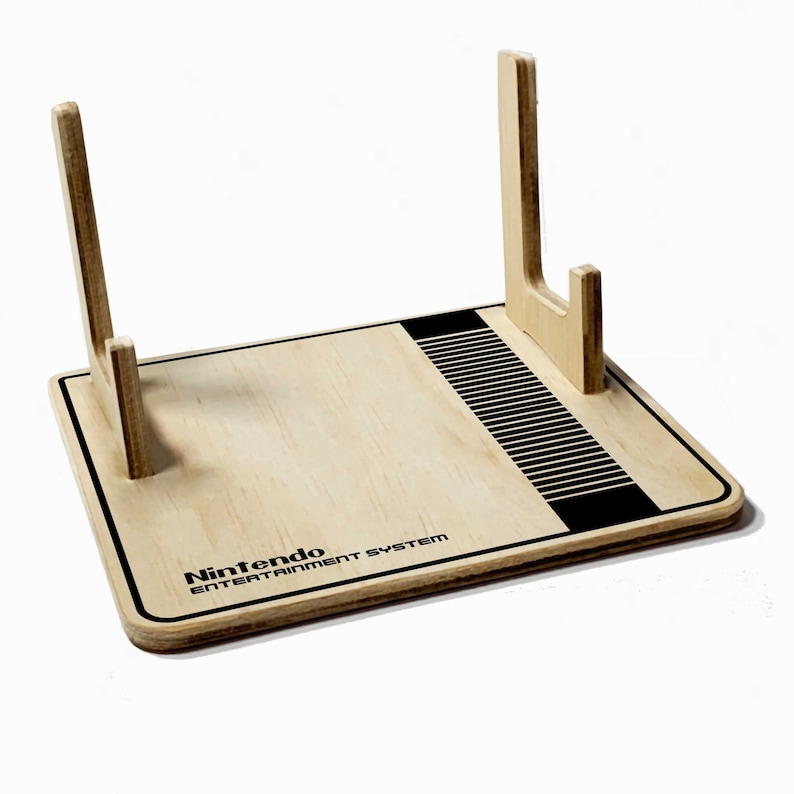 May include: A wooden Nintendo Entertainment System game console stand. The stand is light-colored with black accents, including a design on the right side and the words "Nintendo Entertainment System" printed in black. Two upright supports are on the stand.