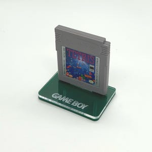 May include: A green acrylic Game Boy stand with the text "GAME BOY" engraved on the front. A gray Game Boy cartridge is displayed on the stand. The cartridge has the text "Nintendo GAME BOY" and "TETRIS" on the label.